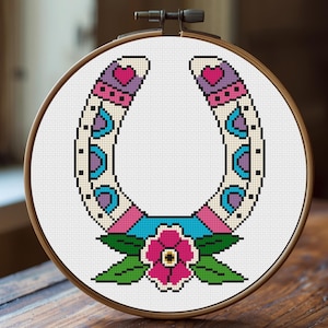 A cross-stitch design of a horseshoe in a wooden embroidery hoop. The horseshoe is decorated with pink, purple, blue, and green patterns, and a pink flower with green leaves is at the bottom. The background is white.