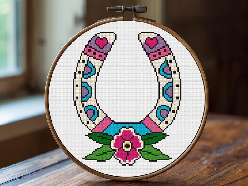A cross-stitch design of a horseshoe in a wooden embroidery hoop. The horseshoe is decorated with pink, purple, blue, and green patterns, and a pink flower with green leaves is at the bottom. The background is white.