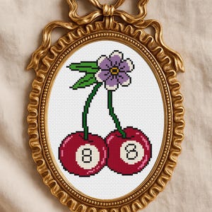 May include: A framed cross-stitch featuring two cherries with the number 8 on each, topped with a purple flower and green leaves. The artwork is set in an ornate gold oval frame with a bow at the top.