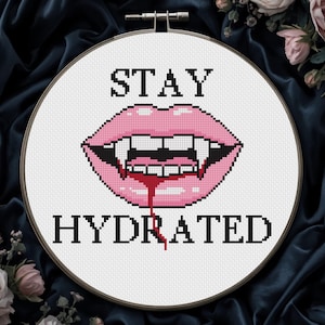 May include: Cross-stitch embroidery hoop with the words "STAY HYDRATED" above and below a pixelated image of pink lips with fangs and dripping blood. The embroidery is set against a dark blue fabric background.