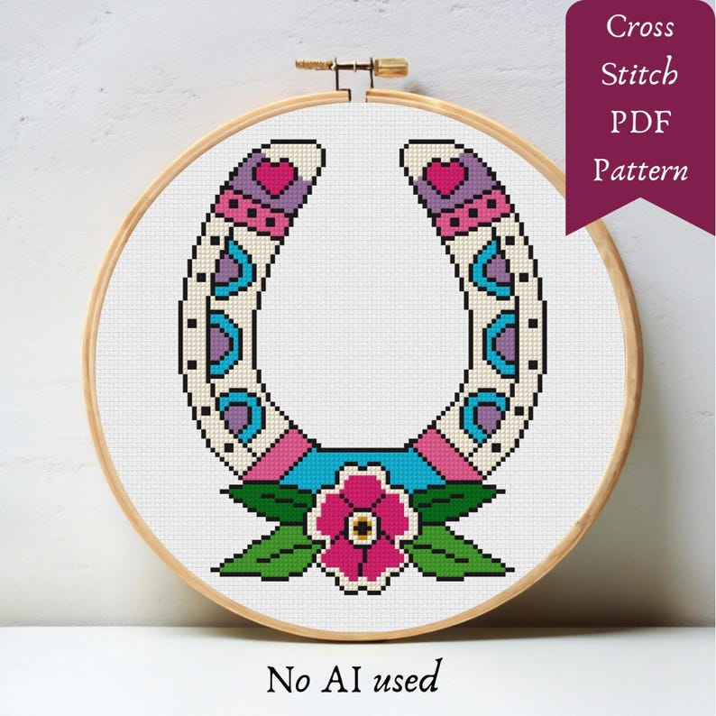 A cross-stitch pattern of a horseshoe with a floral design, displayed in a wooden embroidery hoop. The horseshoe is white with pink, purple, and blue accents. A pink flower and green leaves are at the base. Text on the image reads "Cross Stitch PDF Pattern" and "No AI used."