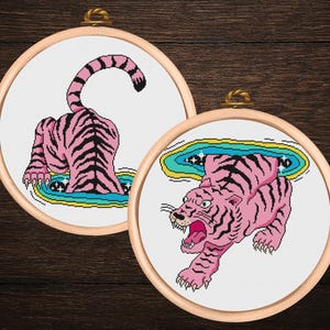 Puede incluir: Dos aros de bordado de punto de cruz con tigres rosas con rayas negras. Uno muestra la parte trasera del tigre, el otro un tigre rugiente que emerge de un portal. Los aros están sobre un fondo de madera oscura.