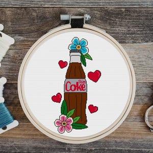 May include: A cross-stitch design of a Diet Coke bottle with red hearts and flowers. The bottle is brown with a silver label and the words "Diet Coke". The design is in a wooden embroidery hoop.