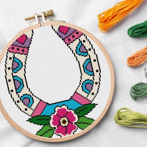 A completed cross-stitch project in a wooden hoop. The design features a colorful horseshoe with a pink flower and green leaves at the bottom. Embroidery floss in orange, green, and other colors are in the background.