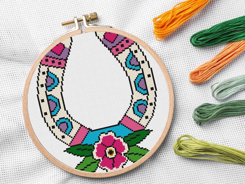 A completed cross-stitch project in a wooden hoop. The design features a colorful horseshoe with a pink flower and green leaves at the bottom. Embroidery floss in orange, green, and other colors are in the background.