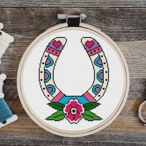 A cross-stitch design of a horseshoe in a wooden embroidery hoop. The horseshoe is decorated with pink, purple, blue, and cream-colored patterns, and a pink flower with green leaves is at the bottom. Embroidery floss and scissors are in the background.