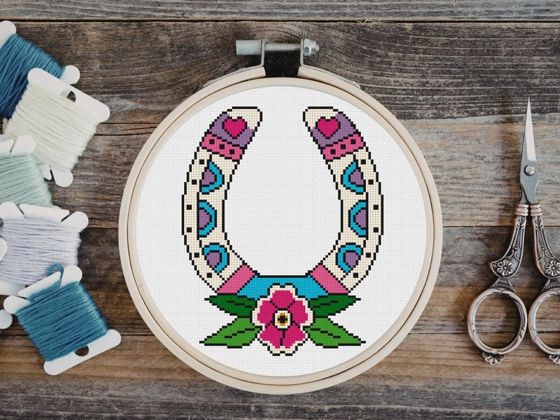 A cross-stitch design of a horseshoe in a wooden embroidery hoop. The horseshoe is decorated with pink, purple, blue, and cream-colored patterns, and a pink flower with green leaves is at the bottom. Embroidery floss and scissors are in the background.