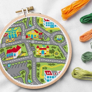 May include: A cross-stitch embroidery hoop with a colourful town map design, including roads, buildings, and a roundabout. The embroidery is set in a wooden hoop. Bright embroidery floss in orange, green, and yellow are arranged nearby.