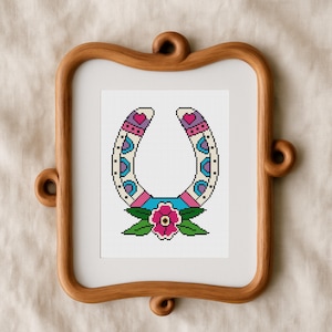 A framed cross-stitch design featuring a horseshoe with a floral motif. The horseshoe is white with pink, blue, and purple accents. A pink flower with green leaves is at the base. The frame is made of wood.