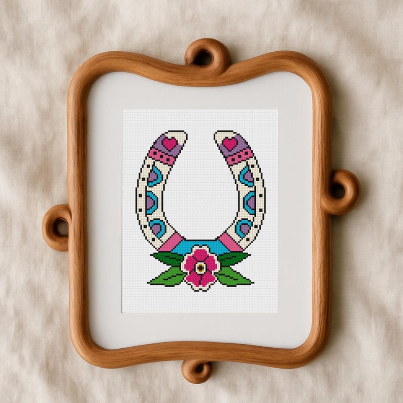 A framed cross-stitch design featuring a horseshoe with a floral motif. The horseshoe is white with pink, blue, and purple accents. A pink flower with green leaves is at the base. The frame is made of wood.