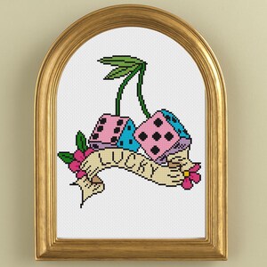 May include: Cross-stitch artwork in a gold frame. The design features two dice in pink and blue, with the word "LUCKY" on a banner. A cherry stem and leaves are above the dice, with a pink flower on the bottom.