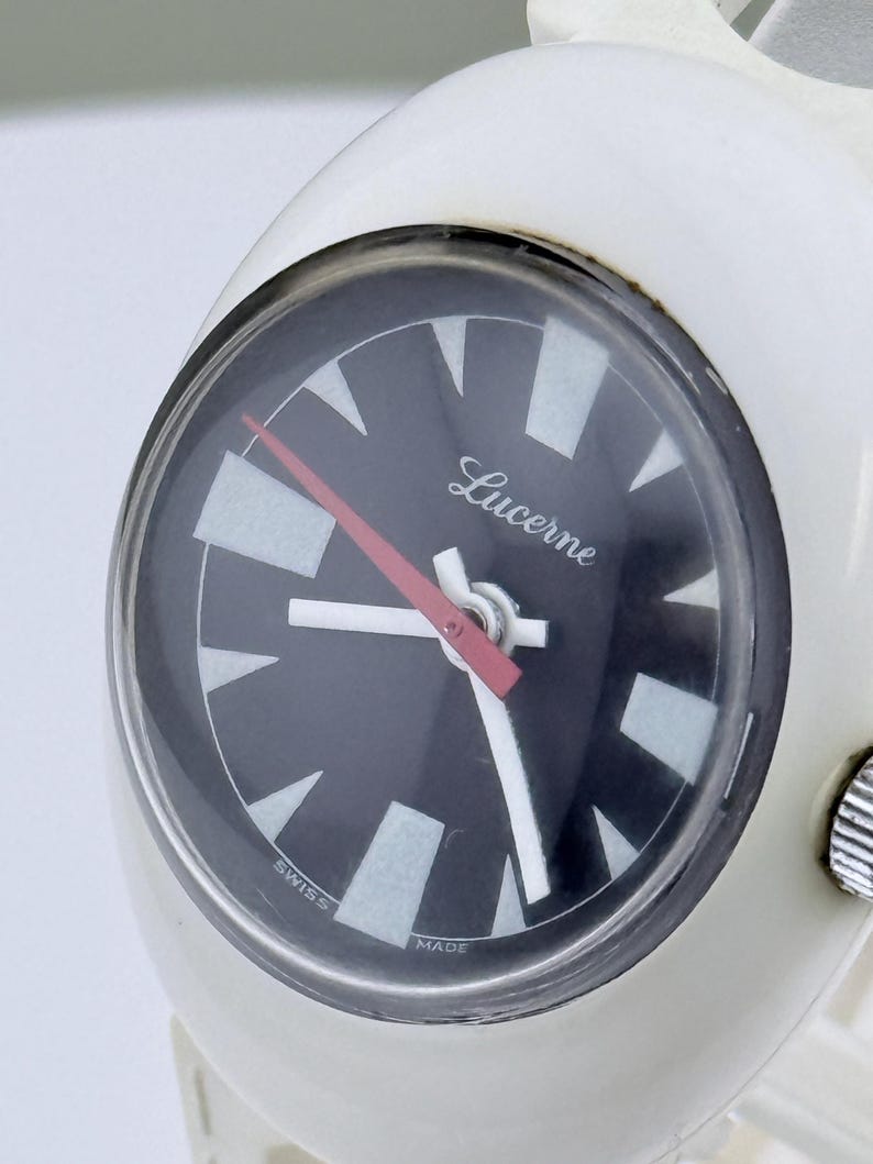 Puede incluir: Un reloj de pulsera Lucerne blanco con esfera negra y agujas blancas y rojas. La esfera del reloj tiene marcadores de hora triangulares blancos y las palabras "Lucerne", "Swiss" y "Made".