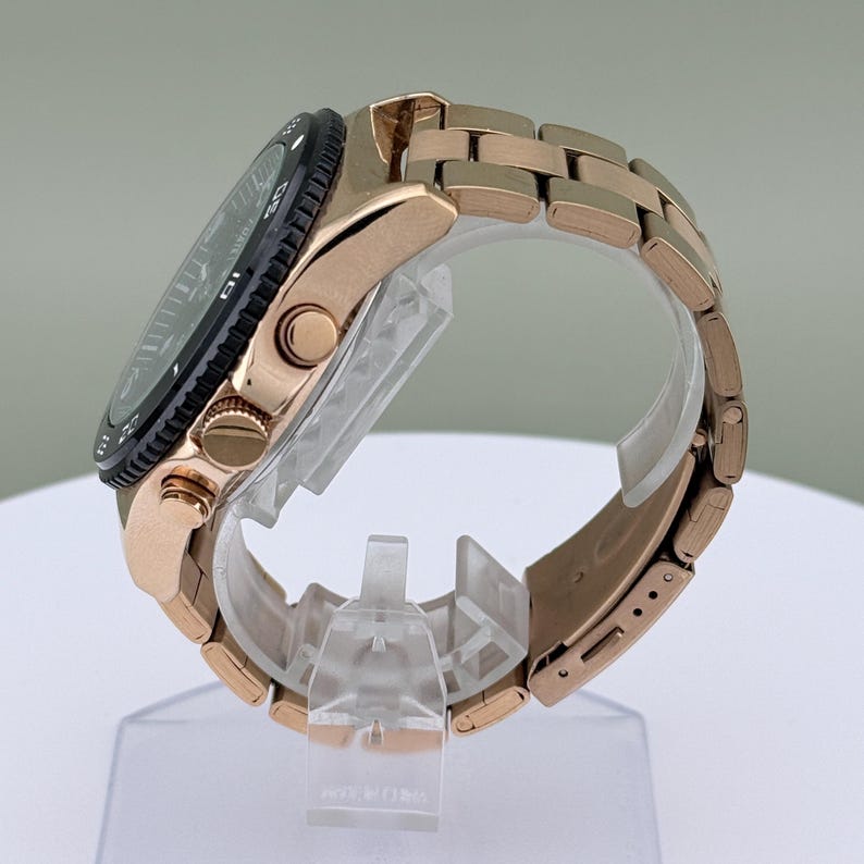 K&ouml;nnte beinhalten: Ros&eacute;goldfarbene Armbanduhr mit schwarzer L&uuml;nette und passendem Gliederarmband. Die Uhr wird auf einem transparenten St&auml;nder pr&auml;sentiert, der ihr Seitenprofil zeigt. Das Zifferblatt der Uhr ist dunkel, mit sichtbaren Markierungen und Zeigern.