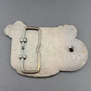 May include: A vintage metal belt buckle with a rectangular frame and a decorative, irregular shape. The buckle has a tarnished, aged appearance, with visible scratches and wear. The design includes a stylized letter "J" on the left side.