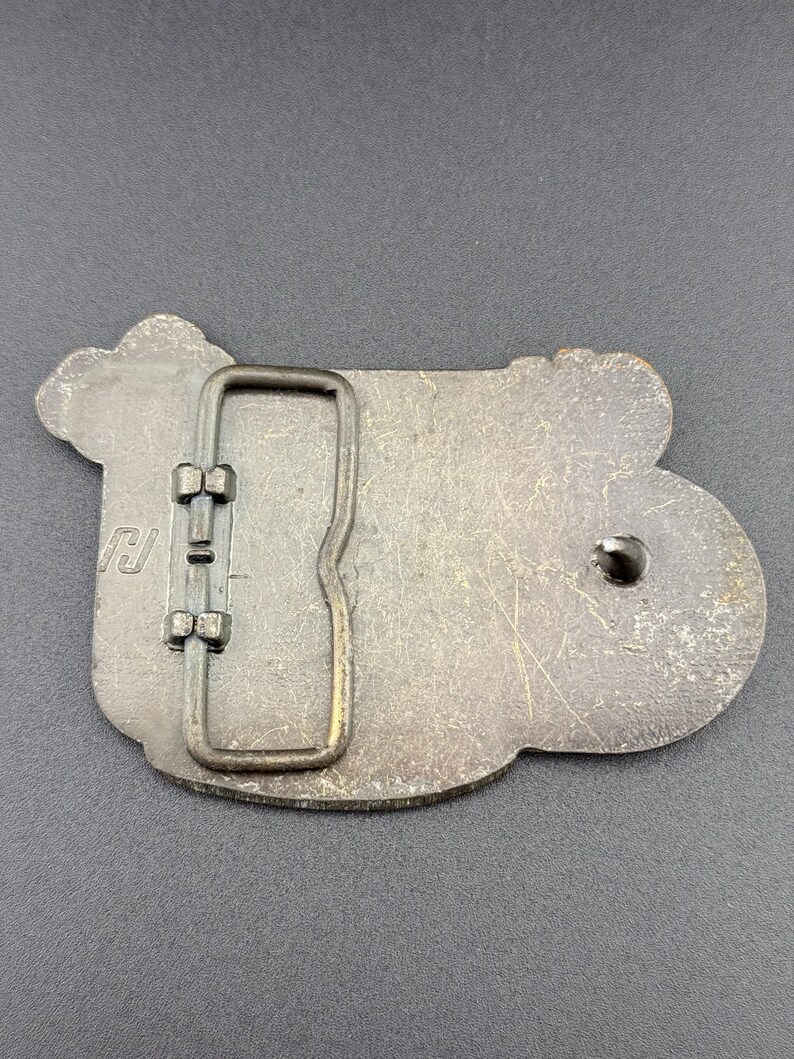 May include: A vintage metal belt buckle with a rectangular frame and a decorative, irregular shape. The buckle has a tarnished, aged appearance, with visible scratches and wear. The design includes a stylized letter "J" on the left side.