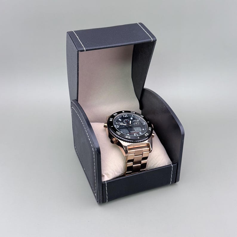 K&ouml;nnte beinhalten: Eine ros&eacute;goldene Armbanduhr mit schwarzem Zifferblatt und L&uuml;nette liegt in einer schwarzen Uhrenbox aus Kunstleder mit wei&szlig;en N&auml;hten. Die Uhr hat ein Metallarmband und wird auf einem cremefarbenen Kissen pr&auml;sentiert.