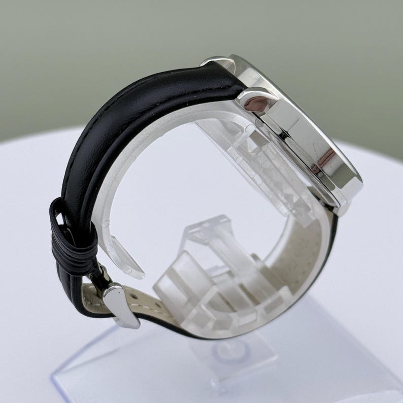 May include: A silver wristwatch with a black leather band. The watch face is round and the band is secured with a silver buckle. The watch is displayed on a clear plastic stand.