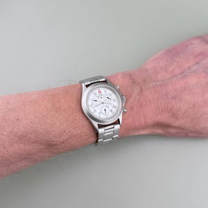 Puede incluir: Un reloj de pulsera plateado con esfera blanca y m&uacute;ltiples subesferas. El reloj tiene una correa de acero inoxidable y un logotipo rojo y blanco. El reloj est&aacute; en la mu&ntilde;eca de una persona.