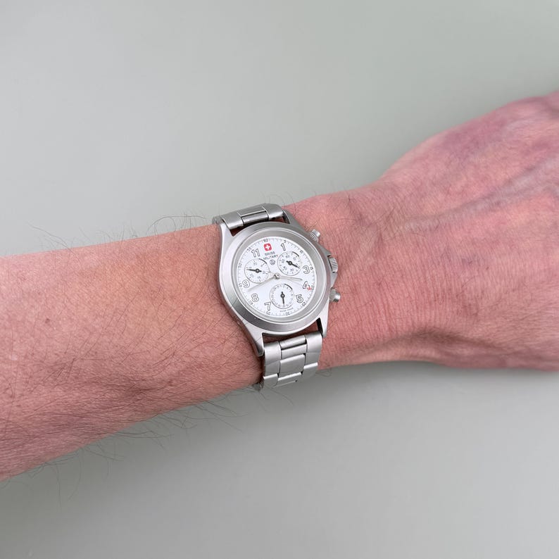 Puede incluir: Un reloj de pulsera plateado con esfera blanca y m&uacute;ltiples subesferas. El reloj tiene una correa de acero inoxidable y un logotipo rojo y blanco. El reloj est&aacute; en la mu&ntilde;eca de una persona.