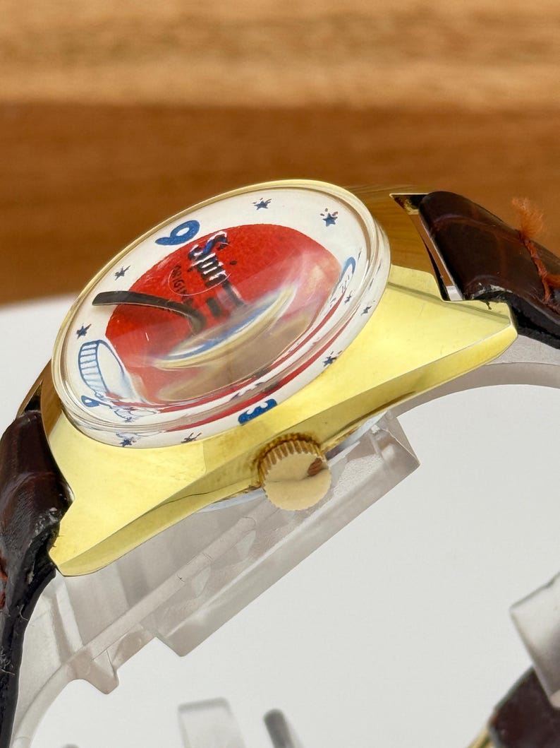 May include: A vintage wristwatch with a gold-toned case and a brown leather strap. The watch face features a red and white design with the text "Spin" and blue star accents. The watch has a gold crown.