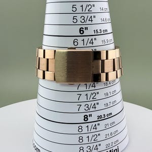 K&ouml;nnte beinhalten: Ein ros&eacute;goldenes Uhrenarmband wird auf einem wei&szlig;-schwarzen Ringma&szlig; angezeigt. Das Uhrenarmband besteht aus rechteckigen Gliedern und hat einen rechteckigen Verschluss. Das Ringma&szlig; hat Zoll- und Zentimetermessungen.