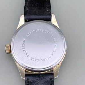 K&ouml;nnte beinhalten: Nahaufnahme einer Vintage-Armbanduhr mit schwarzem Lederarmband. Die Uhr hat ein goldfarbenes Metallgeh&auml;use und einen Edelstahlboden. Auf der R&uuml;ckseite der Uhr stehen die Worte "Stainless Steel Back", "Base Metal Case" und "Dustproof".