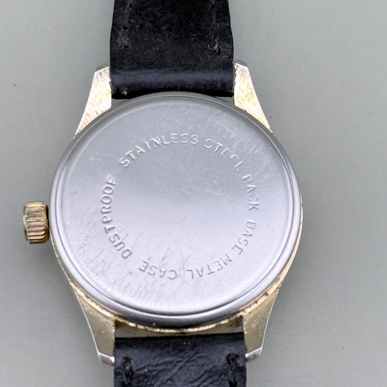 K&ouml;nnte beinhalten: Nahaufnahme einer Vintage-Armbanduhr mit schwarzem Lederarmband. Die Uhr hat ein goldfarbenes Metallgeh&auml;use und einen Edelstahlboden. Auf der R&uuml;ckseite der Uhr stehen die Worte "Stainless Steel Back", "Base Metal Case" und "Dustproof".