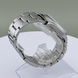Puede incluir: Un reloj de pulsera plateado con una correa de acero inoxidable. El reloj se muestra sobre un soporte de pl&aacute;stico transparente. La correa est&aacute; compuesta por eslabones rectangulares. La esfera del reloj no es visible.