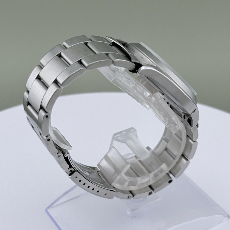 Puede incluir: Un reloj de pulsera plateado con una correa de acero inoxidable. El reloj se muestra sobre un soporte de pl&aacute;stico transparente. La correa est&aacute; compuesta por eslabones rectangulares. La esfera del reloj no es visible.