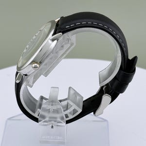 May include: A silver-toned wristwatch with a black leather band. The watch face is visible, and the band is secured with a silver buckle. The band has white stitching along the edges. The watch is displayed on a clear plastic stand.