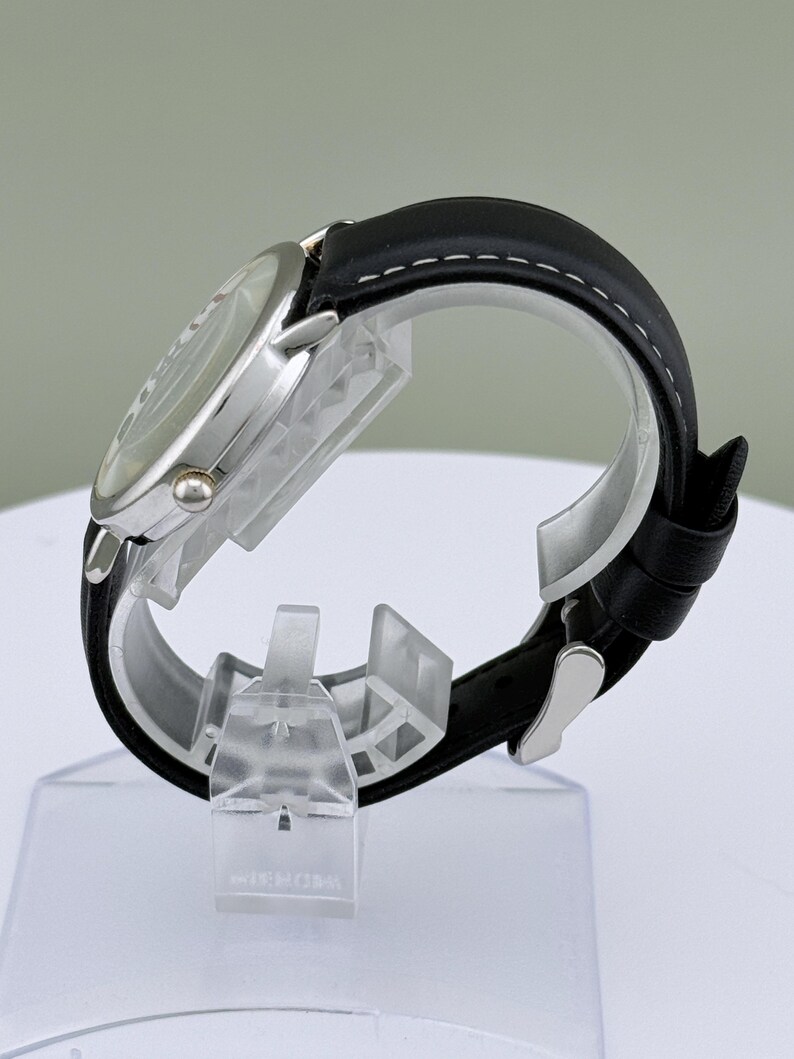 May include: A silver-toned wristwatch with a black leather band. The watch face is visible, and the band is secured with a silver buckle. The band has white stitching along the edges. The watch is displayed on a clear plastic stand.