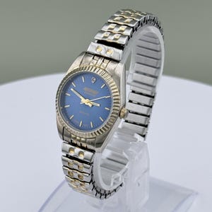 May include: A silver and gold-toned quartz watch with a blue face. The watch features a gold bezel and hands, with the brand name "Nile River Diamond" displayed. The watch band is a silver and gold-toned metal link style.