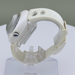 Puede incluir: Un reloj de pulsera blanco con esfera redonda y correa blanca. El reloj tiene una esfera plateada y una correa blanca texturizada con agujeros de ajuste. El reloj se exhibe sobre un soporte transparente.