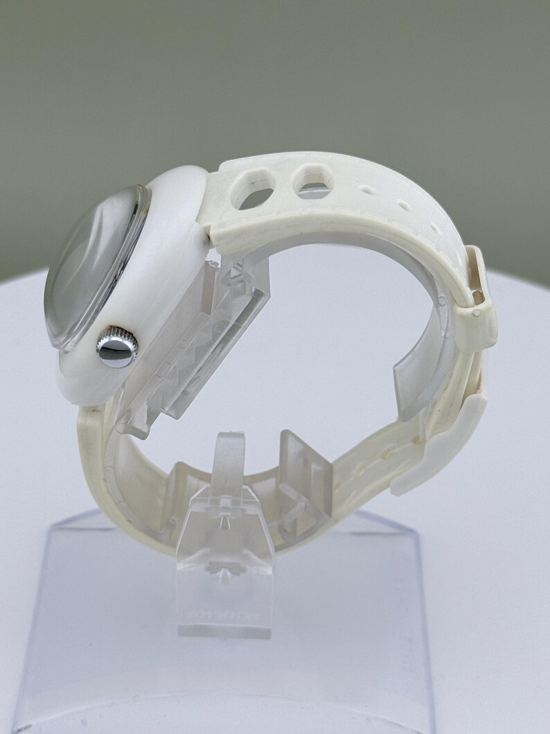 Puede incluir: Un reloj de pulsera blanco con esfera redonda y correa blanca. El reloj tiene una esfera plateada y una correa blanca texturizada con agujeros de ajuste. El reloj se exhibe sobre un soporte transparente.