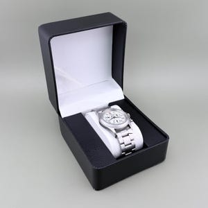 Puede incluir: Un reloj de pulsera plateado con esfera blanca y correa plateada descansa dentro de una caja de reloj negra con interior blanco. El reloj tiene m&uacute;ltiples esferas y una correa de acero inoxidable. La caja est&aacute; abierta, revelando el reloj.