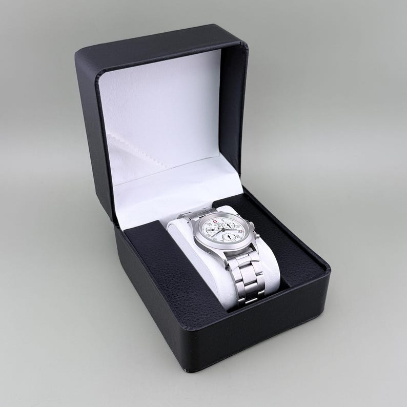 Puede incluir: Un reloj de pulsera plateado con esfera blanca y correa plateada descansa dentro de una caja de reloj negra con interior blanco. El reloj tiene m&uacute;ltiples esferas y una correa de acero inoxidable. La caja est&aacute; abierta, revelando el reloj.