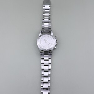Puede incluir: Un reloj de pulsera de acero inoxidable plateado con una correa de eslabones y una esfera redonda. El reloj tiene una funci&oacute;n de cron&oacute;grafo. El reloj se muestra sobre un fondo gris. La correa del reloj est&aacute; hecha de m&uacute;ltiples eslabones rectangulares.