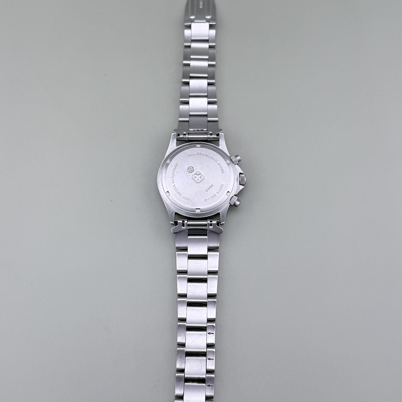 Puede incluir: Un reloj de pulsera de acero inoxidable plateado con una correa de eslabones y una esfera redonda. El reloj tiene una funci&oacute;n de cron&oacute;grafo. El reloj se muestra sobre un fondo gris. La correa del reloj est&aacute; hecha de m&uacute;ltiples eslabones rectangulares.