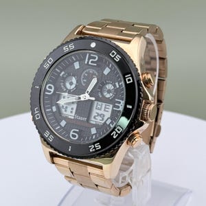 K&ouml;nnte beinhalten: Eine goldfarbene Armbanduhr mit schwarzer L&uuml;nette und dunkelgrauem Zifferblatt. Die Uhr verf&uuml;gt &uuml;ber mehrere Zifferbl&auml;tter, digitale Anzeigen und ein goldfarbenes Metallarmband. Das Zifferblatt zeigt die Marke "Stauer".