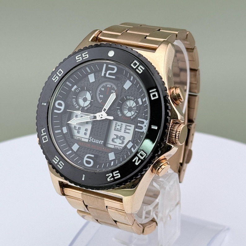 K&ouml;nnte beinhalten: Eine goldfarbene Armbanduhr mit schwarzer L&uuml;nette und dunkelgrauem Zifferblatt. Die Uhr verf&uuml;gt &uuml;ber mehrere Zifferbl&auml;tter, digitale Anzeigen und ein goldfarbenes Metallarmband. Das Zifferblatt zeigt die Marke "Stauer".