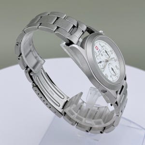 Puede incluir: Un reloj Swiss Military de acero inoxidable con esfera blanca y m&uacute;ltiples subesferas. El reloj tiene una correa plateada y un logotipo de cruz roja. El reloj se muestra sobre un soporte transparente.