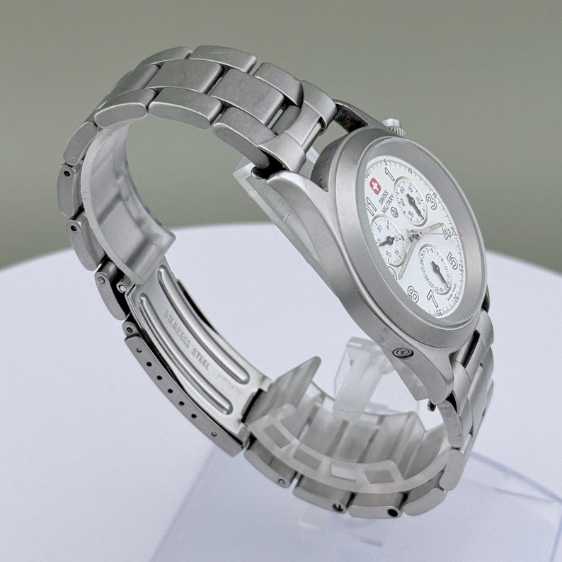 Puede incluir: Un reloj Swiss Military de acero inoxidable con esfera blanca y m&uacute;ltiples subesferas. El reloj tiene una correa plateada y un logotipo de cruz roja. El reloj se muestra sobre un soporte transparente.