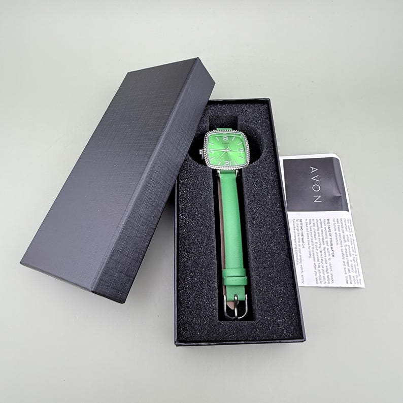 May include: A green wristwatch with a square face and a green leather strap is presented in a black box. The watch face has a silver bezel. A card with the word "AVON" is also visible. The watch is a stylish accessory.