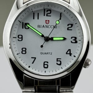 Pu&ograve; includere: Orologio da polso argentato con quadrante bianco, numeri neri e lancette luminose. Il quadrante mostra il marchio "BIANCCHI" e la parola "QUARTZ". L'orologio ha un cinturino argentato e una corona laterale.