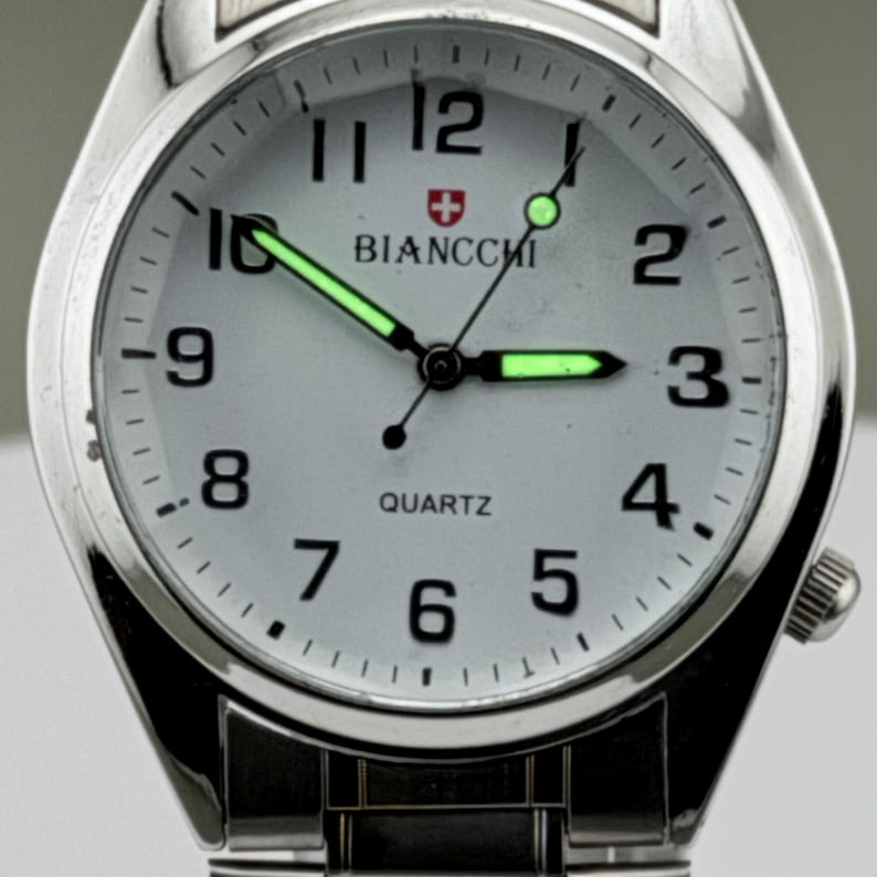 Pu&ograve; includere: Orologio da polso argentato con quadrante bianco, numeri neri e lancette luminose. Il quadrante mostra il marchio "BIANCCHI" e la parola "QUARTZ". L'orologio ha un cinturino argentato e una corona laterale.