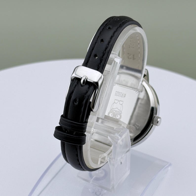 May include: A silver-toned wristwatch with a black leather band. The watch has a round face and a buckle closure. The back of the watch has the text "#R3525" and a small graphic. The watch is displayed on a clear stand.