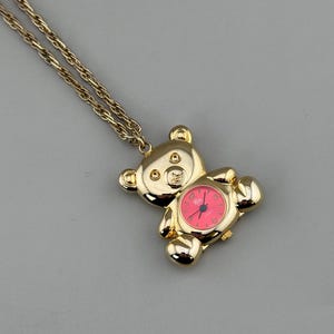May include: A gold-colored teddy bear pendant watch with a pink watch face and gold chain. The watch face has black numbers and hands. The bear has a detailed design with visible ears, arms, and legs. The chain is a delicate, twisted link style.
