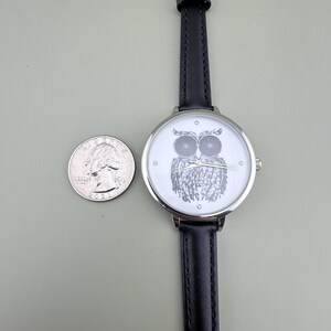 May include: A silver-toned wristwatch with a black leather band and a white face featuring a detailed owl illustration. A U.S. quarter is next to the watch for size comparison. The watch has a classic design.