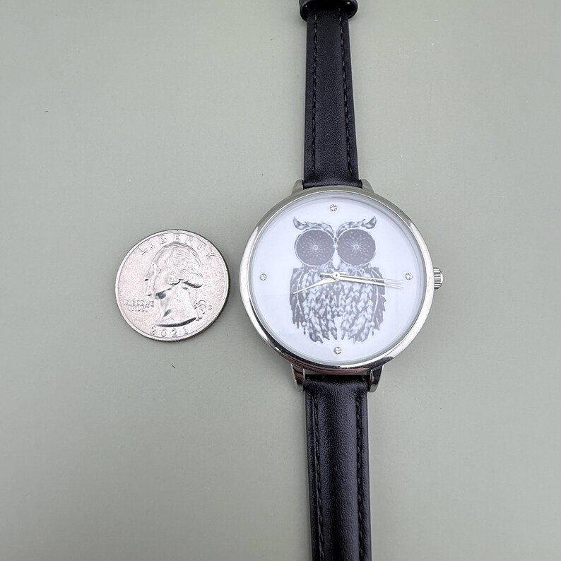 May include: A silver-toned wristwatch with a black leather band and a white face featuring a detailed owl illustration. A U.S. quarter is next to the watch for size comparison. The watch has a classic design.