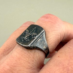 May include: A silver-toned ring with a rectangular face featuring a fleur-de-lis emblem. The ring has a worn, antique appearance, with a detailed design on the face. The ring is a piece of jewelry.